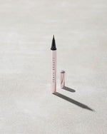 The Flyliner Longwear Liquid Eyeliner standing upright on a light grey concrete surface. The eyeliner is open, revealing its bold black tip, while its exterior features a sleek light metallic pink finish.