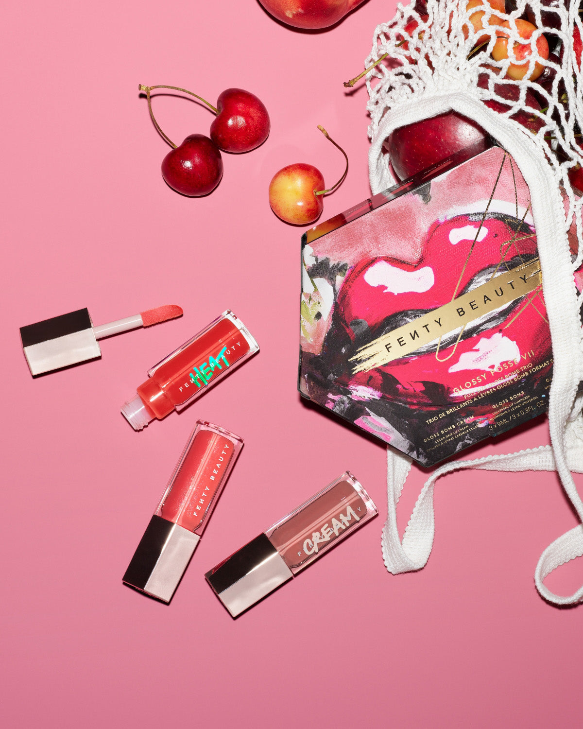 Gloss Bomb Heat Universal Lip Luminizer + Plumper in shade 'Hot Cherry' and cherry fruits in a grocery bag on a pink background.